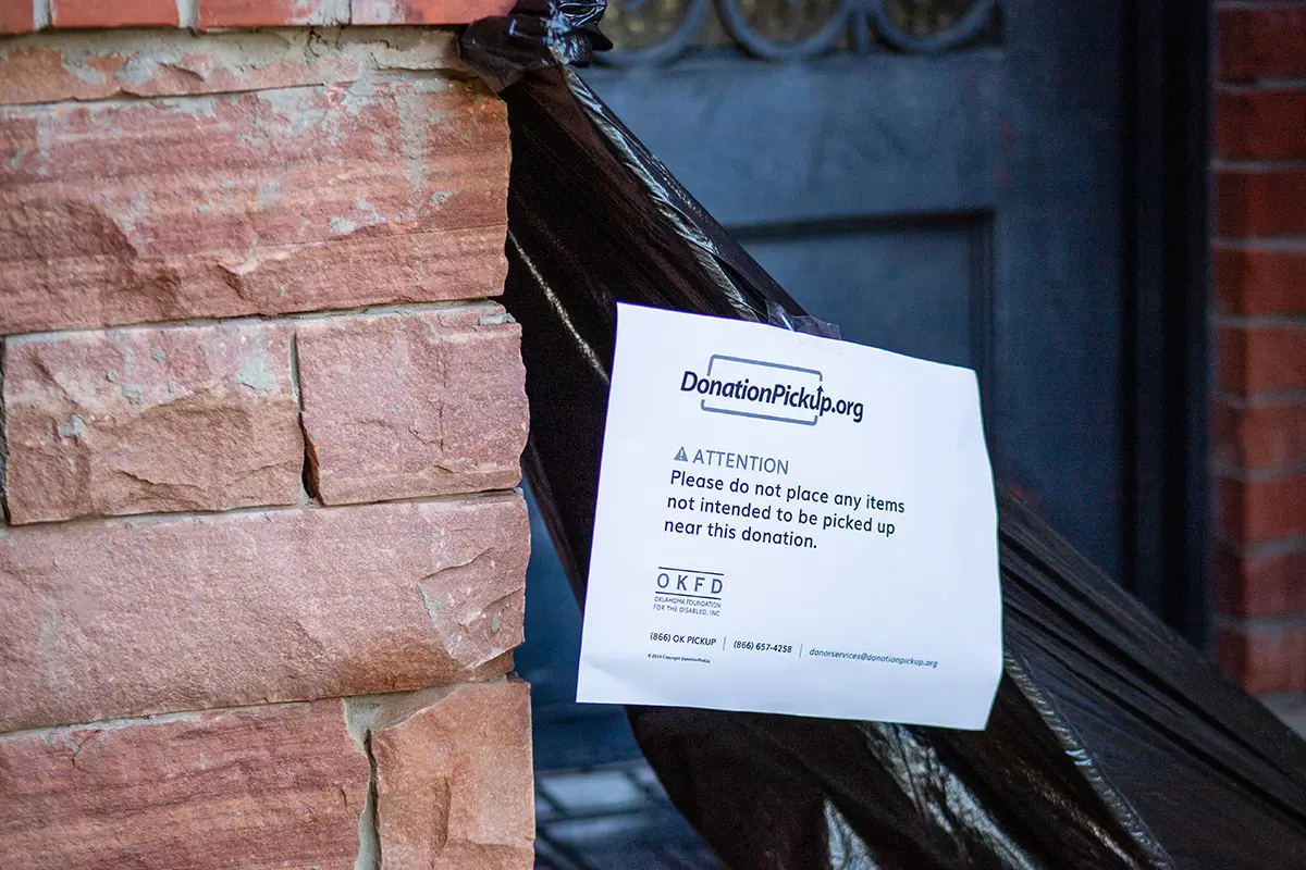A close-up of a black trash bag placed outdoors against a brick wall. A white paper sign from DonationPickup.org is taped to it, asking not to place other items near it.