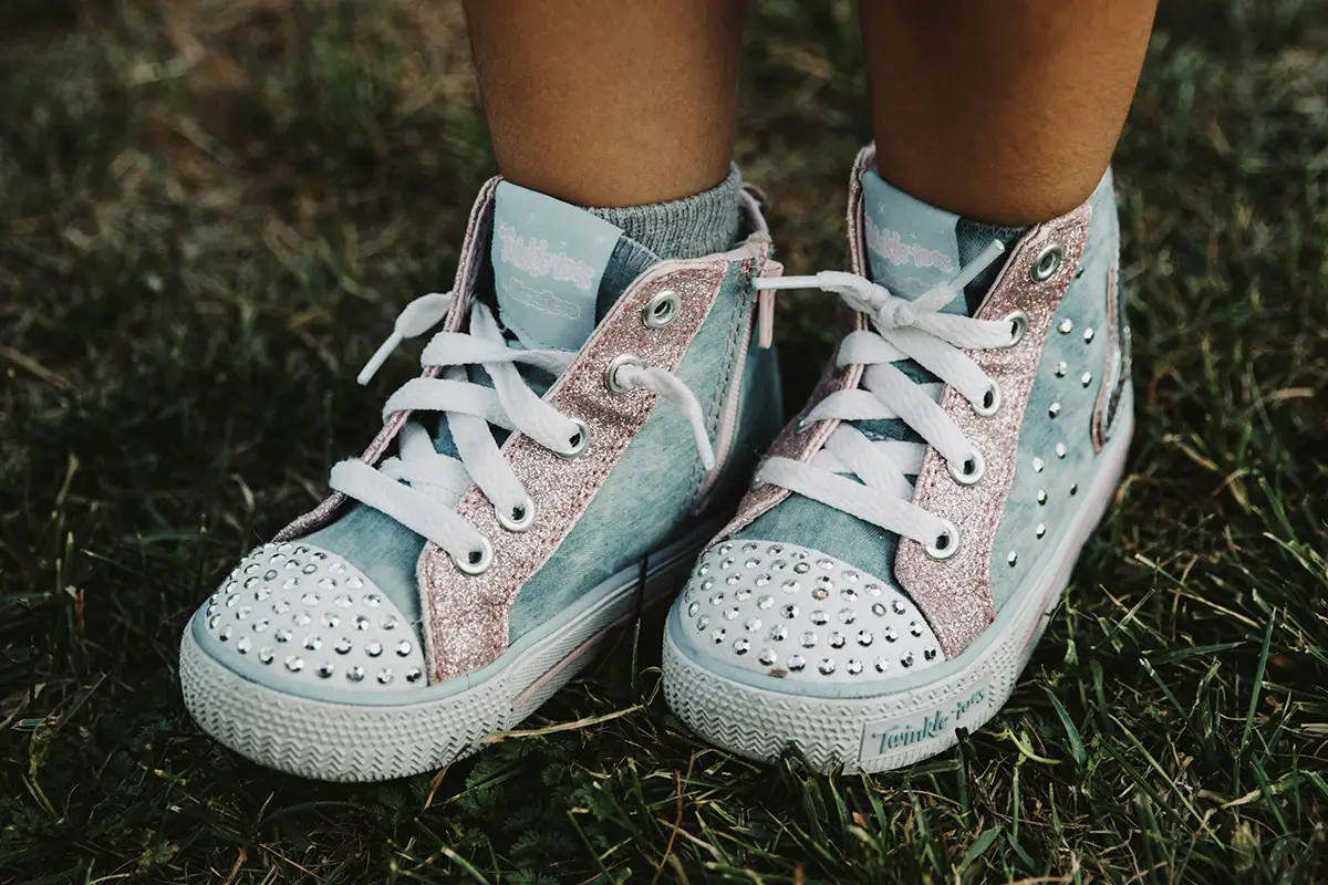 An eye-level shot of a child wearing light blue and sparkly pink high-top Skechers Twinkle Toes sneakers while standing in the grass.